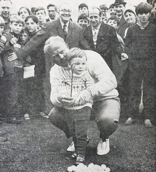 a crowd looks on as a crouching Jack Nicklaus helps a young boy play a shot during the NCR clinic in 1968