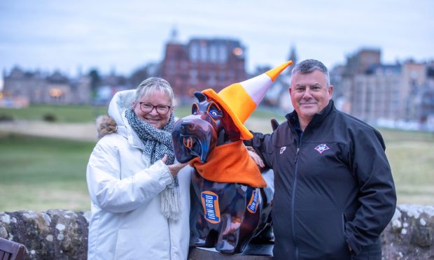 Image shows: Kym and James Shields with Bru Dog, a model of a Scottie Dog with Iron Bru logos and a fabric traffic cone hat. The couple are standing near the Old Course in St Andrews with the town in the background.