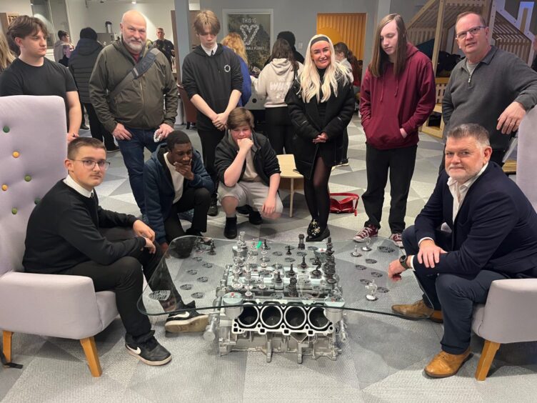 Man and teenage boy at glass-topped chess table made from Porsche engine, watched by group of people at Y Centre 