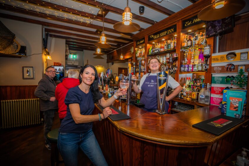 Gayle sits at the bar at at The Old Ship Inn, posing for a picture with the bartender. 