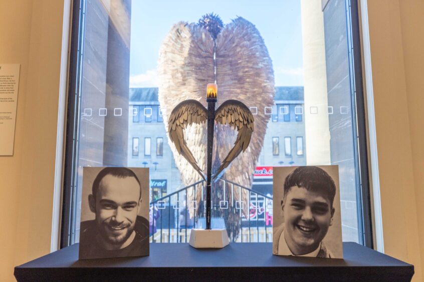 Photos of Cameron Rae and Barry Dixon on a table inside Perth Museum, with knife angel visible, from the back, through window onto street outside.