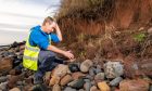 Warden Stuart Cook kneels at the skeleton find site with coastal erosion visible in the background.