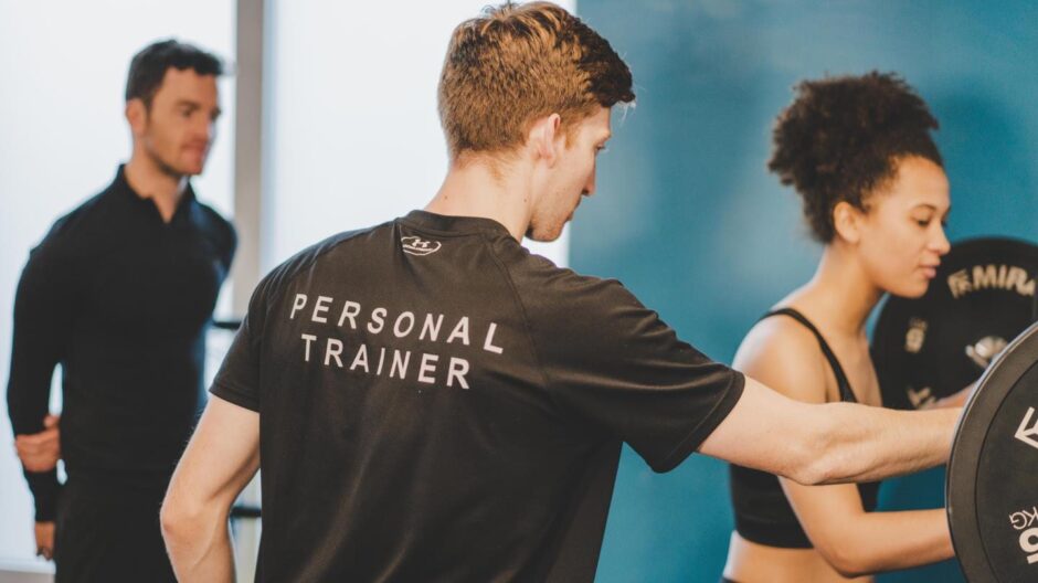 two men, one with personal trainer on the back of his t-shirt, help a woman to lift weights