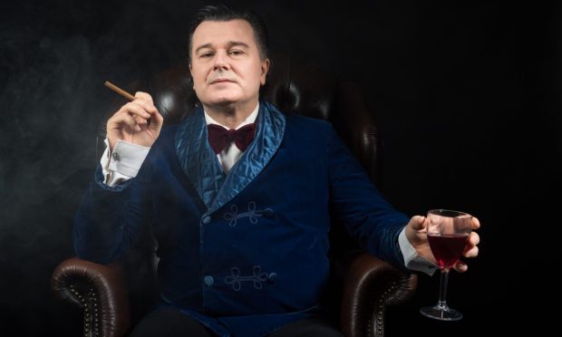 Image shows: Gerard Logan in his one-man show Night Terrors. Gerard is dressed in a dark blue velvet smoking jacket with white shirt and burgandy velvet bow-tie. He is sitting in a wing-backed chair and holding a slim cigar in his right hand and a glass of red wine in his left hand.