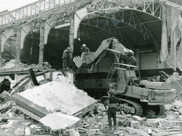 a man looks on as a digger gets to work on the half-demolished building