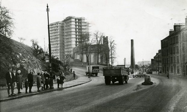 People at the bus stop on Lochee Road, at the foot of Dudhope Terrace, in January 1965, with a high-rise block inn the distance and lorries on the road.