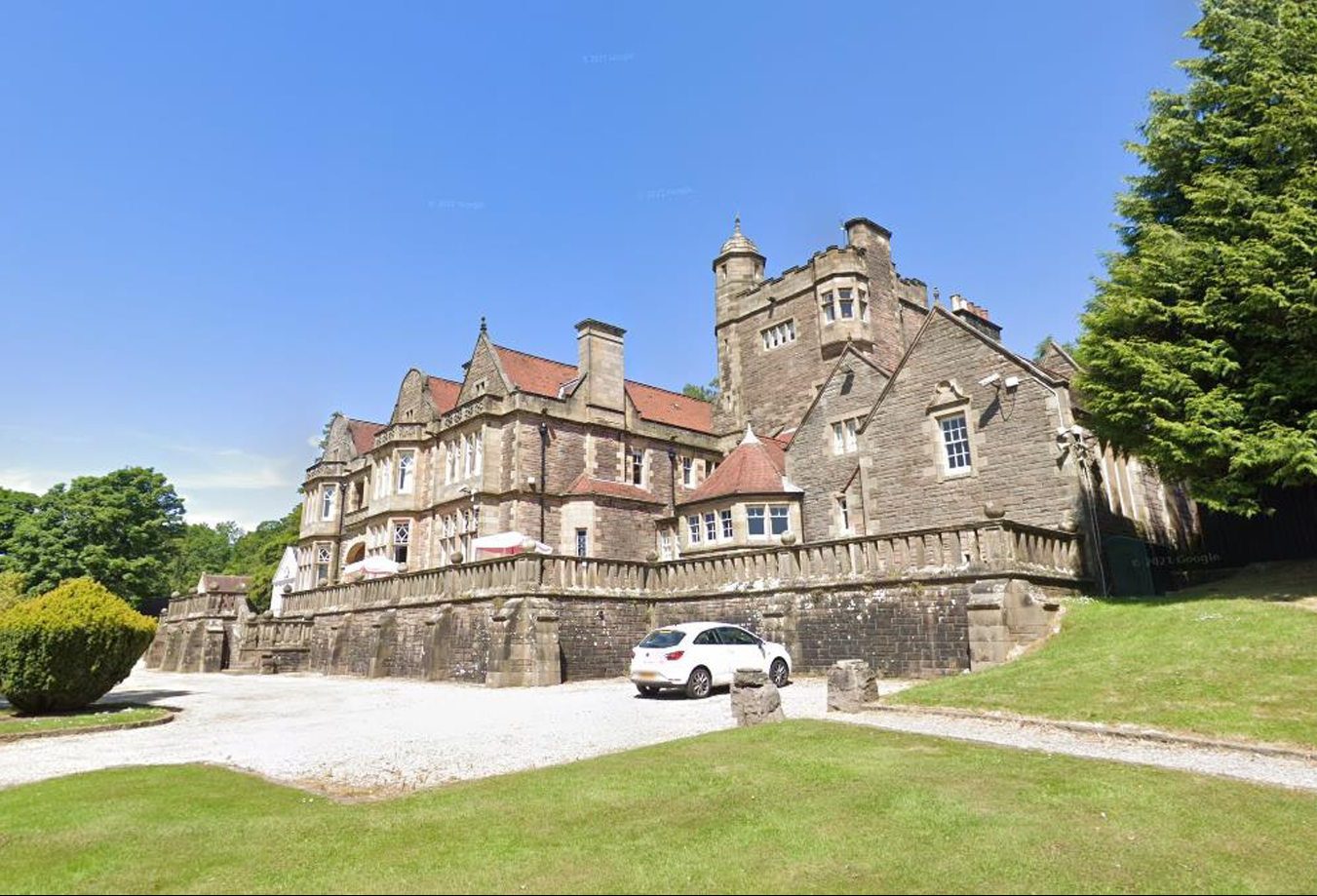 The castle-like, stone Inglewood House hotel and spa, with a red roof, is in the background, with lawns in the foreground. The sky is blue and there are green bushes to each side of the photo