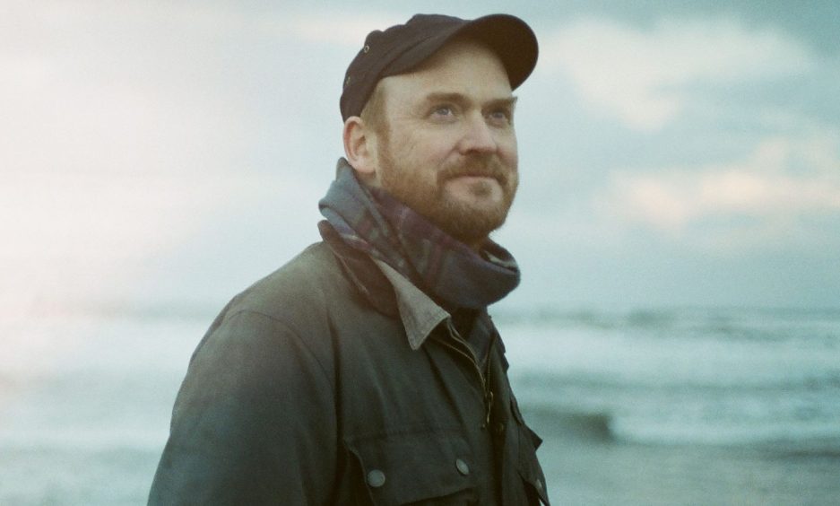 Image shows: Fife musician and writer James Yorkston. James is pictured outdoors with a wavy sea in the background. He is wearing a black cap and has red hair and a red beard.He is looking up into the sky with a wry smile.