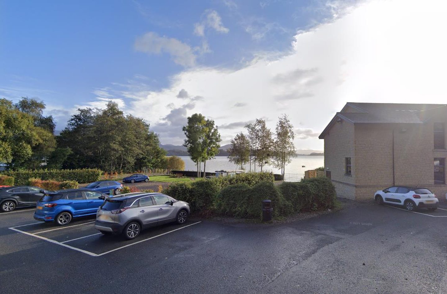 The car park of Lodge on Loch Lomond, with cars parked in the foreground and a nice view of the loch, trees and blue sky in the background. Part of the beige brick hotel building can be seen to the right