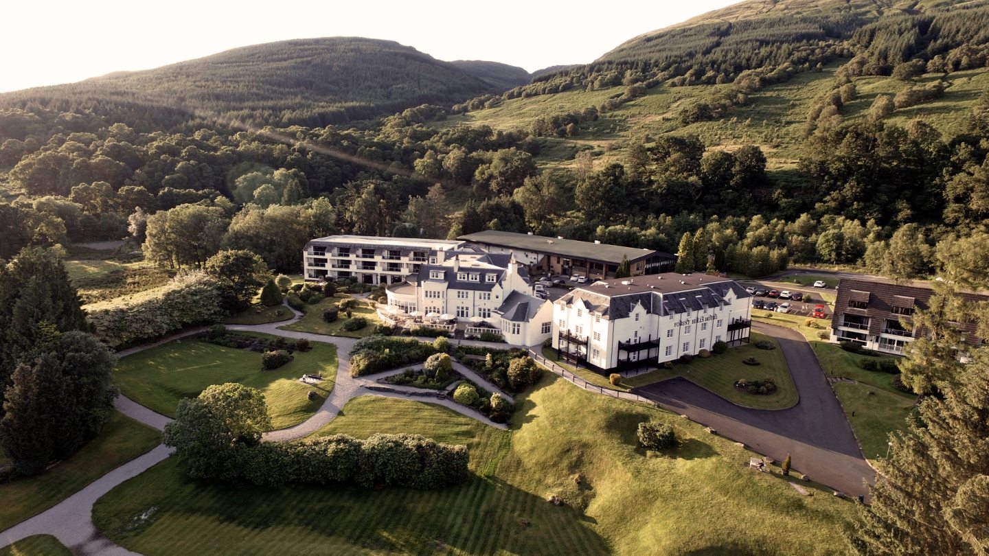 A faraway view of the sprawling Macdonald Forest Hills Hotel and Resort - a while building with black roof, with rolling green hills in the background