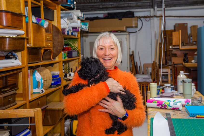 Sarah |Peterson holding small black fluffy dog in her arms with furniture workshop behind