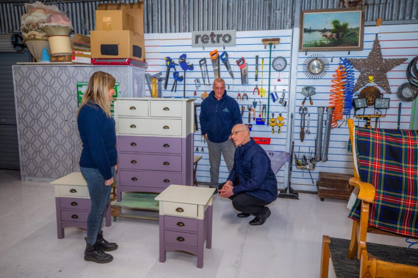 John Swinney kneeling down to look at a set of painted bedroom furniture with wall of tools behind