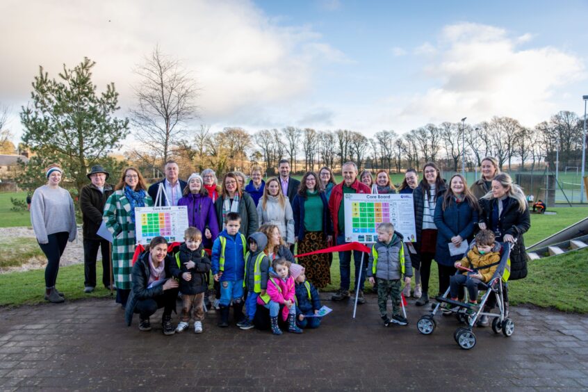 Large group of adults and children standing around two communication boards on easels with park in background