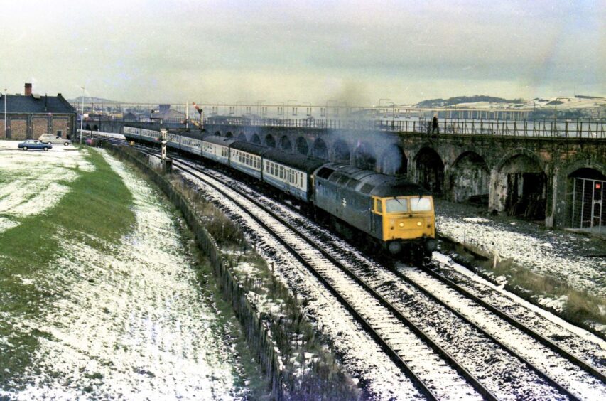 A train goes along tracks beside the River Tay. The Tay Road Bridge can be seen in the distance.