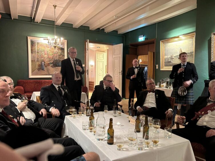 Group of men in kilts and formal wear standing and seated around a table with drinks in front of them