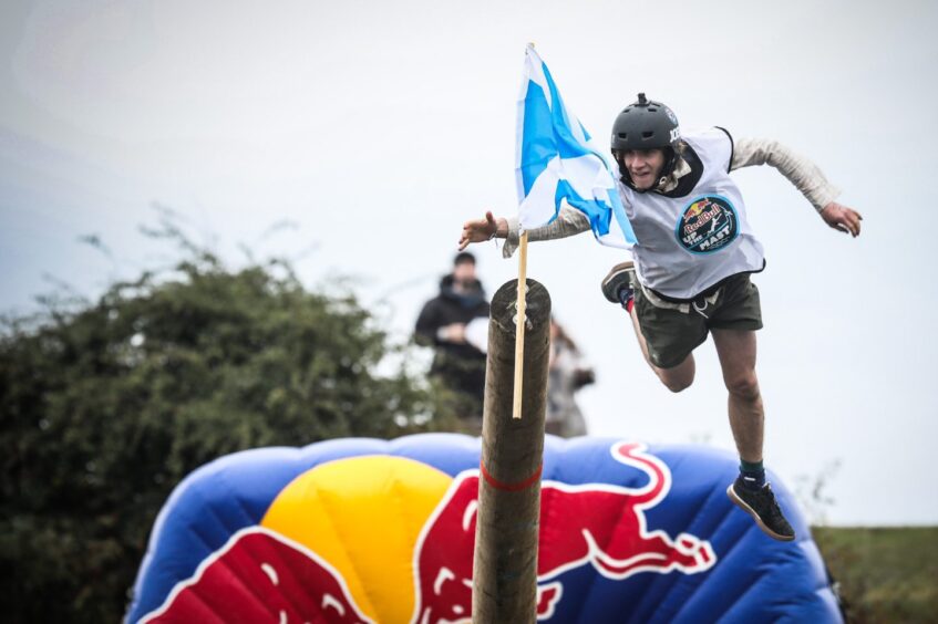 Image shows: a participant in the Red Bull Up the Mast competition in Fife. The person has fallen off the mast and is in mid-air, trying to a Saltire at the end of the mast.