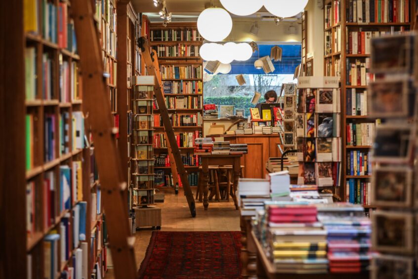 Image shows: an interior view of Topping & Company Booksellers in St Andrews. The long room is lined with bookshelves and tall library ladders. There are tables full of stacks of books in the middle of the space.