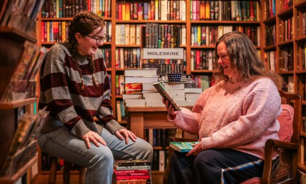 Image shows: feature writer Nora McElhone with bookseller Jen Thomas in Topping & Company, St Andrews. They are seated in a corner of the bookshop surrounded by packed bookcases and with a pile of books stacked on the floor between them.