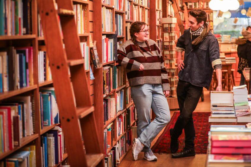 image shows booksellers Jen Thomas and David Thomas (no relation) chatting in Topping & Company Booksellers in St Andrews. Jen is leaning against the bookshelves and is wearing jeans and a striped jumper.David is standing with his legs crossed over and is wearing skinny jeans, a dark jumper and neckscarf.