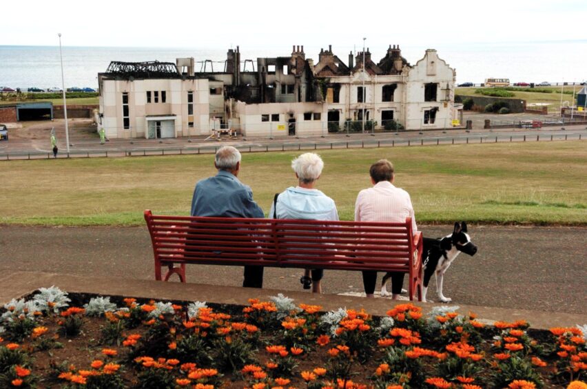 three people sit on a bench looking down at the coastline and the charred remains of Hotel Seaforth