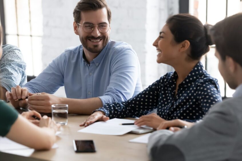 Smiling diverse businesspeople gather at desk in office have fun joke discussing ideas together, happy multiethnic colleagues laugh negotiate brainstorming at meeting in boardroom