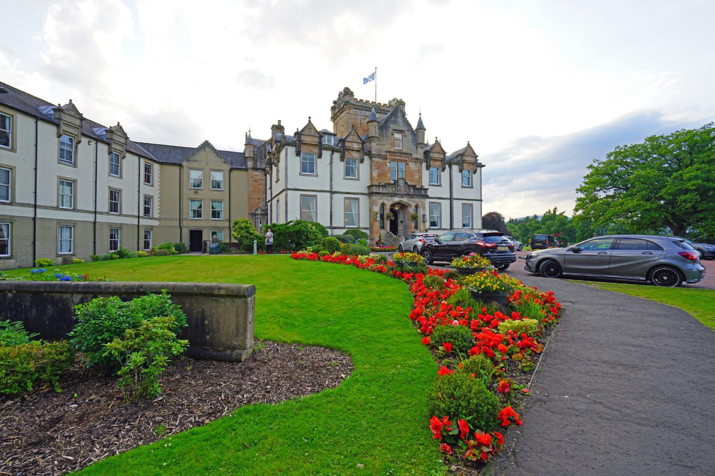 The castle-like sandstone and white exterior of Cameron House hotel, with turrets and a flagpole, flying a Saltire. Some cars are parked outside the hotel, and there is a green lawn and attractive red and yellow flowers planted in the foreground