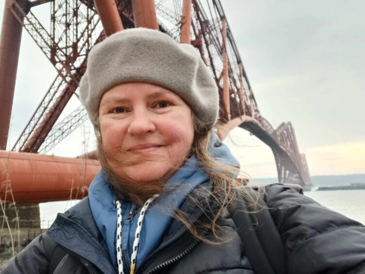 Image shows: A close up of Nora McElhone with the Forth Bridge in the background.Nora is wearing a blue hoodie, black coat and beige beret hat. Her hair is blowing in the wind.