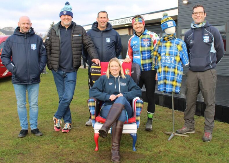 Jayne Strachan seated on chair with group of men and mannequin wearing Doddie Weir foundation shirt behind her