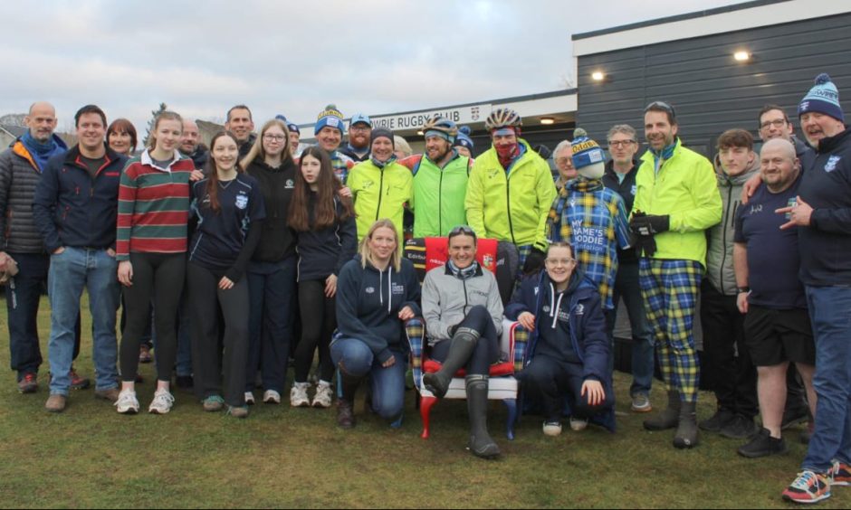 Group of cyclists and supporters grouped around woman in arm chair at Blairgowrie Rugby Club