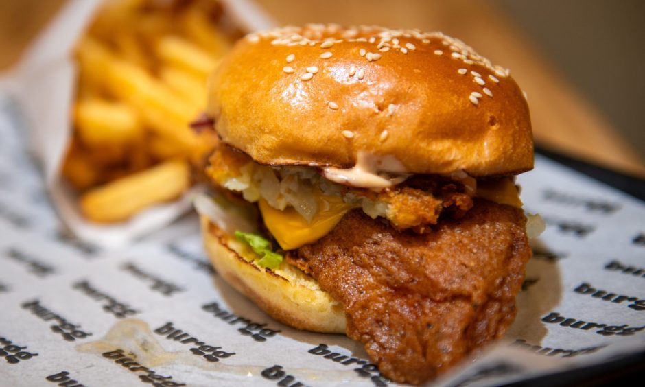 A large fried chicken pattie hanging out of a burger bun with cheese and salad and fires in the background.