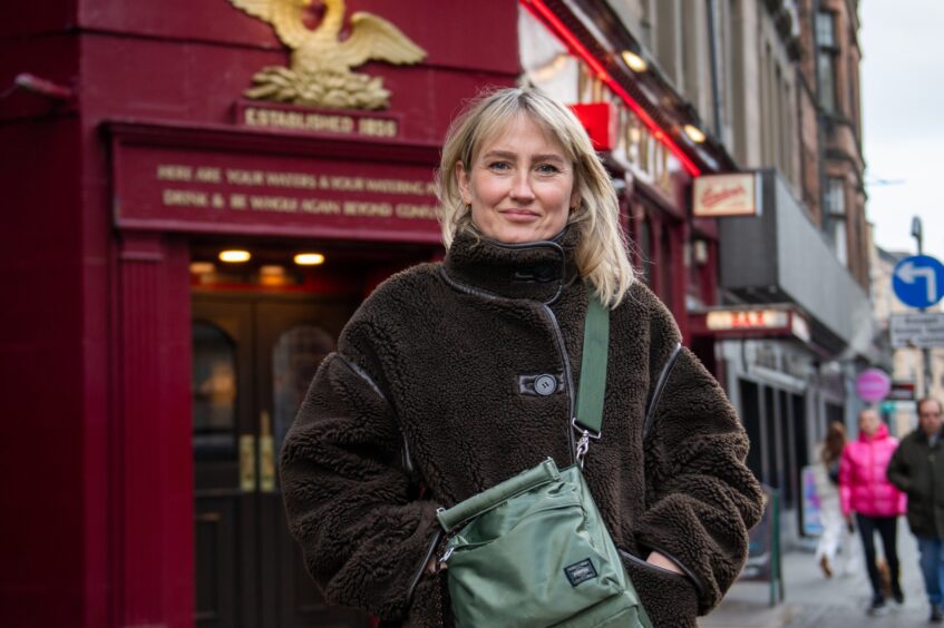 Image shows DC Thomson illustrator standing outside one of her favourite Dundee landmarks, The Phoenix Bar. Kate is standing in front of the red building wearing a brown fluffy coat and a cross-body bag. She has shoulder-length hair with a fringe.