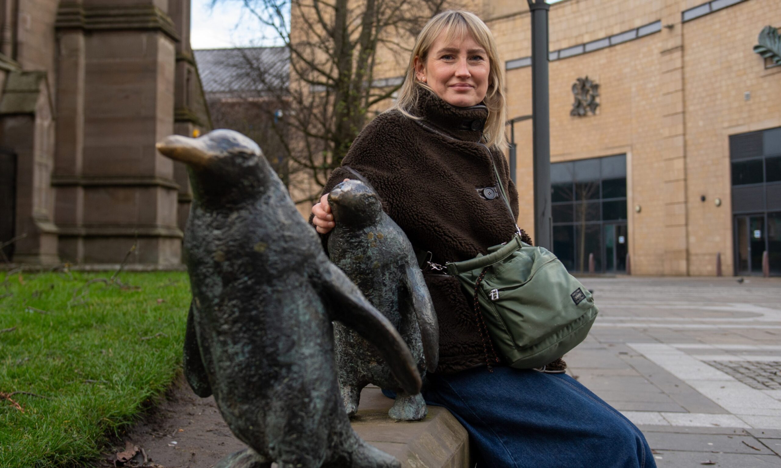 Image shows: Illustrator Kate Benzie with one of her favourite Dundee landmarks: the penguin statues. Kate is sitting amongst the metal statues with her hand on one of the penguins. She is wearing a brown furry jacket and long denim skirt. She has blond hair with a fringe.