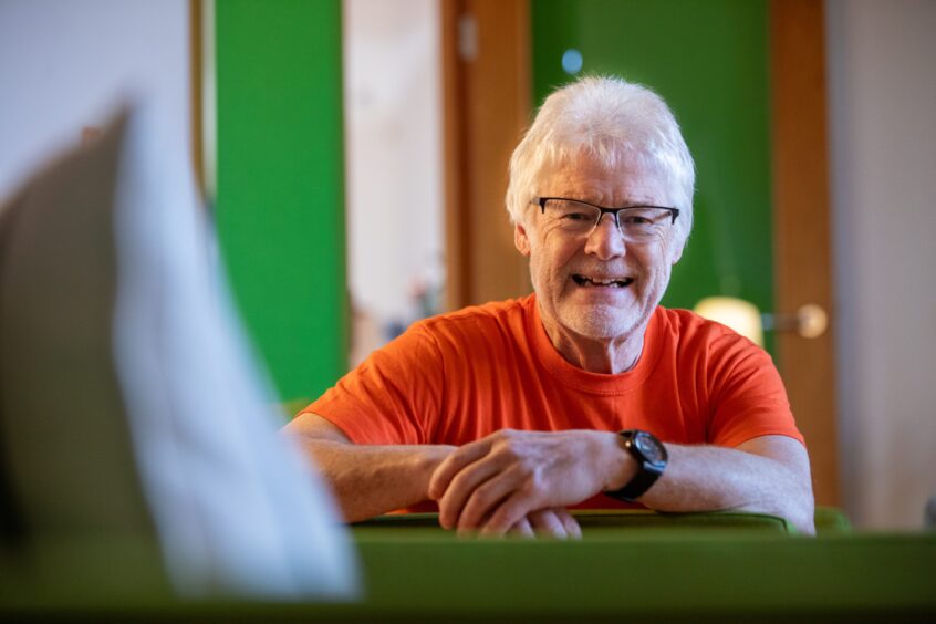 Image shows: Maggie's volunteer Ken Bray. The picture is a close up head and shoulders shot of Ken who is wearing an orange Maggie's t-shirt and has white hair and a white beard and black-framed glasses.