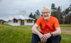 Image shows: Maggie's Centre Dundee volunteer Ken Bray. Ken is sitting on a grass bank outside the Maggie's Centre building. He is resting his hands on his knees and has a warm smile. He has white hair and beard and black glasses.