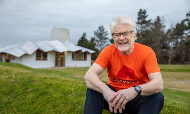 Image shows: Maggie's Centre Dundee volunteer Ken Bray. Ken is sitting on a grass bank outside the Maggie's Centre building. He is resting his hands on his knees and has a warm smile. He has white hair and beard and black glasses.