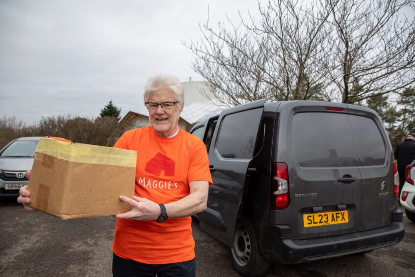 Image shows: Volunteer Ken Bray moving items from his black van to Maggie's Centre Dundee. Ken is wearing a bright orange Maggie's Dundee t-shirt and black trousers and is holding a cardboard box full of leaflets.