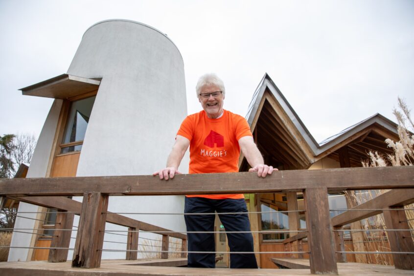 Image shows: Maggie's volunteer Ken Bray standing outside the Dundee Maggie's Centre. Ken is wearing an orange Maggie's Dundee t-shirt and black trousers. He has white hair and beard and dark-rimmed glasses. He is smiling welcomingly.