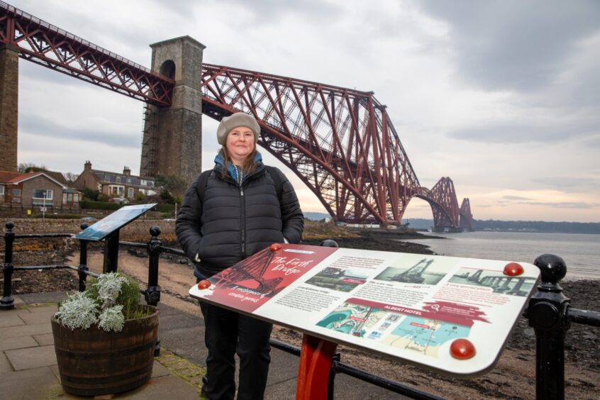 Image shows: Nora McElhone in North Queensferry, standing by an information board for the Forth Bridges Trail. She is wearing a black jacket and beige hat and had her hands in her pockets. The red Forth Bridge dominates the background of the image.