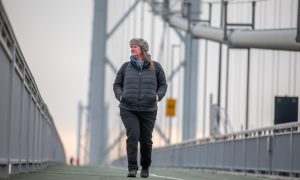 Image shows: feature writer Nora McElhone walking along the Forth Road Bridge. She is wearing dark trousers, a black jacket and beret-style hat and is looking out over the sea. The struts and wires of the bridge can be seen in the background. The sky is grey and overcast.