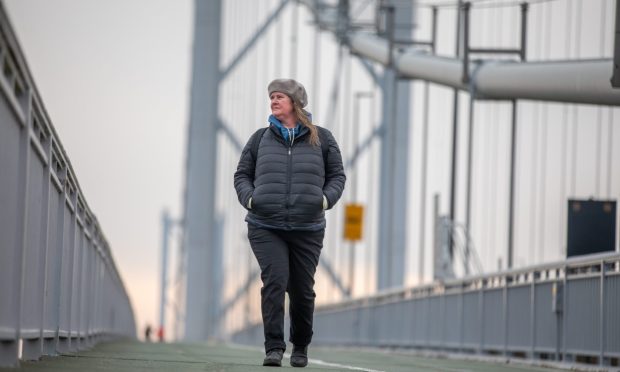 Image shows: feature writer Nora McElhone walking along the Forth Road Bridge. She is wearing dark trousers, a black jacket and beret-style hat and is looking out over the sea. The struts and wires of the bridge can be seen in the background. The sky is grey and overcast.