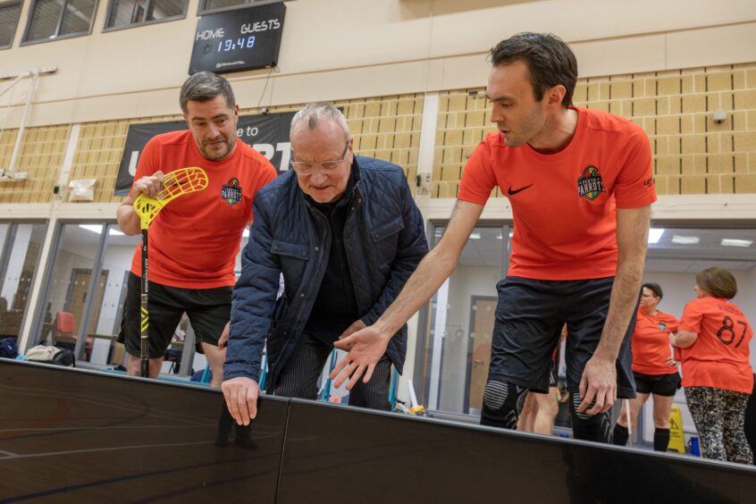 Frazer Robertson, Pete Wishart and Rhuaraidh Fleming leaning over looking at new floorball rink inside gym