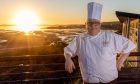 Head chef James McKay stands on the balcony at Horizons restaurant in his full chef's whites and chef's hat with a blazing sunset in the background over the Forth of Firth.