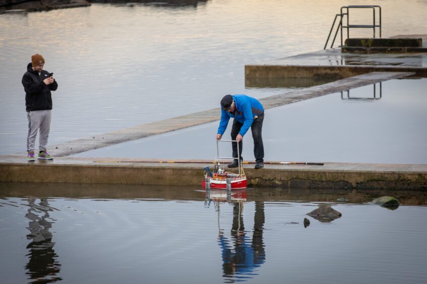 Image shows: Model boat club member taking their boat from the water at Cellardyke Tidal Pool. The calm water reflects the red hull of the boat and blue jacket of the man.