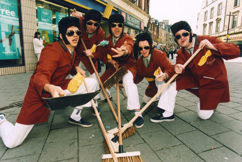 Five men in shades and Elvis wigs with brooms pose for a picture on a Stirling street