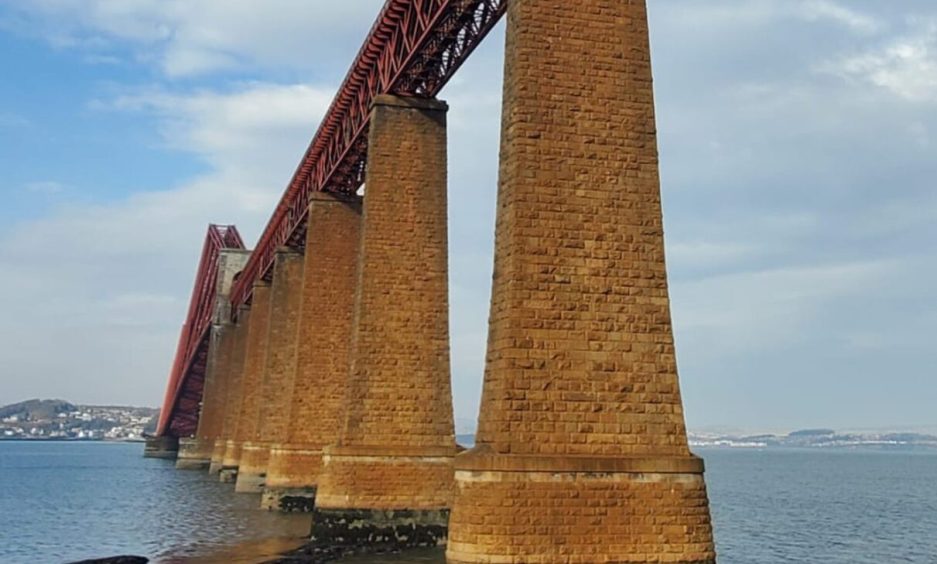 Image shows: The Forth Bridge from South Queensferry. The picture shows the brick bridge supports and red bridge against a blue sky with a few clouds.