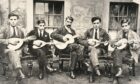 Five men sitting on wooden chairs playing mandolins