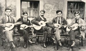 Five men sitting on wooden chairs playing mandolins