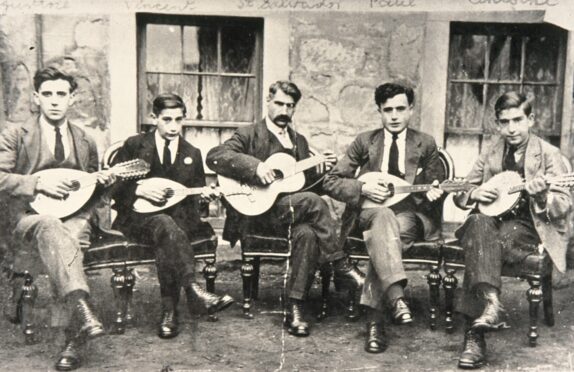 Five men sitting on wooden chairs playing mandolins