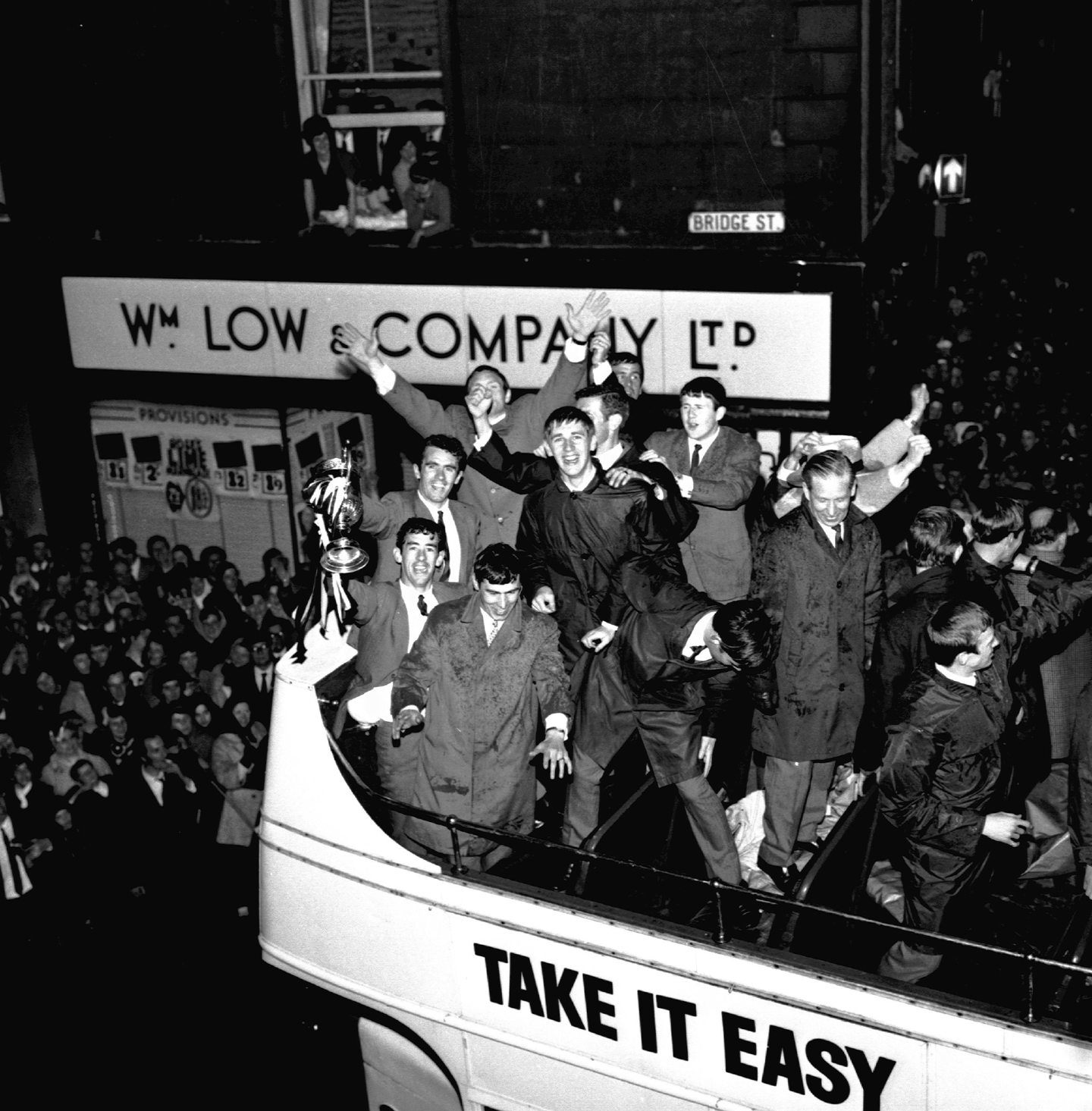 Dunfermline Athletic players showing off the Scottish Cup trophy as the bus makes its way through the crowded city streets in 1961