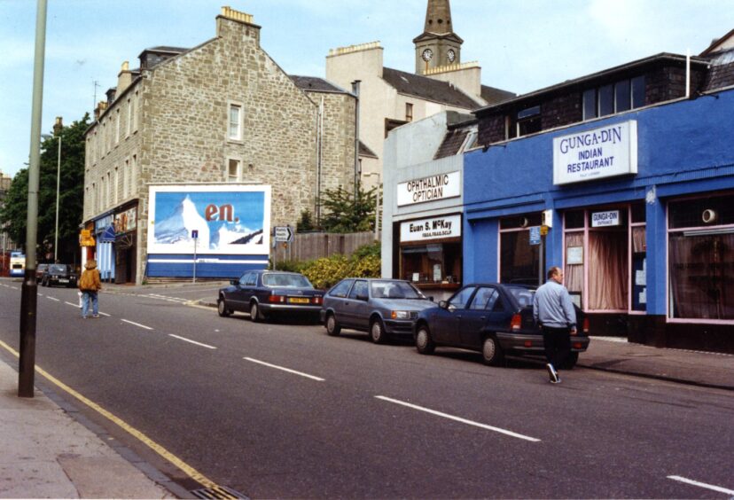 A view of Perth Road in June 1991, showing cars parked on the road and the exterior of McKay optician and Gunga-Din restaurant.